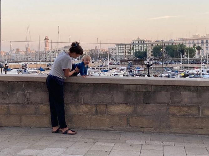 Farbfoto: Aufnahme einer Frau die ein Kind auf einer Mauer hält. Hinter der Mauer befindet sich ein Bootshafen und eine Häuserreihe bei Dämmerung