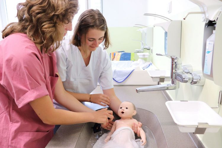 Zwei Studentinnen in medizinischer Kleidung baden eine Neugeborenenpuppe in einem Waschbecken.