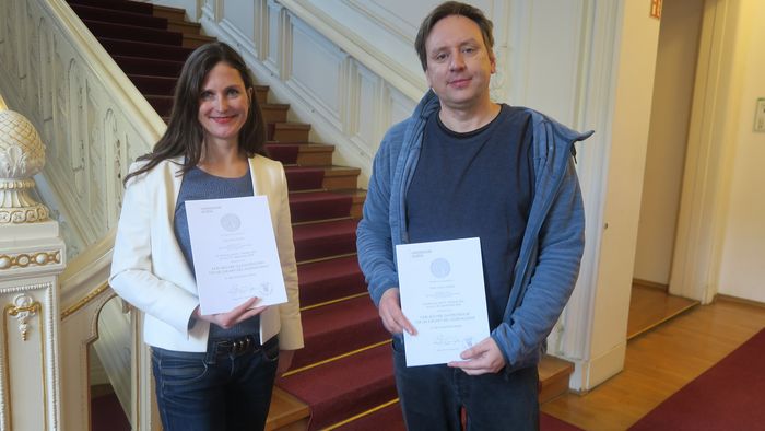 Alina Fichter und Lorenz Matzat zeigen ihre Berufungsurkunden im Foyer des Rektoratsgebäudes.