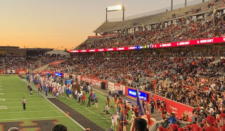 Ein mit Menschen gefülltes Footballstadion in Houston, Texas. Zu sehen ist das Spielfeld und Menschen, die mit dem Rücken zur Kamera stehen.Im Hintergrund geht die Sonne unter.