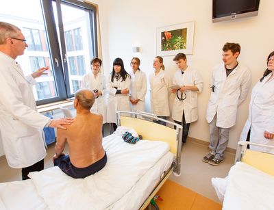 Foto: Studierende in Arztkitteln stehen in einem Patientenzimmer um einen Patient herum der auf einem Bett sitzt, während der Professor etwas erklärt.