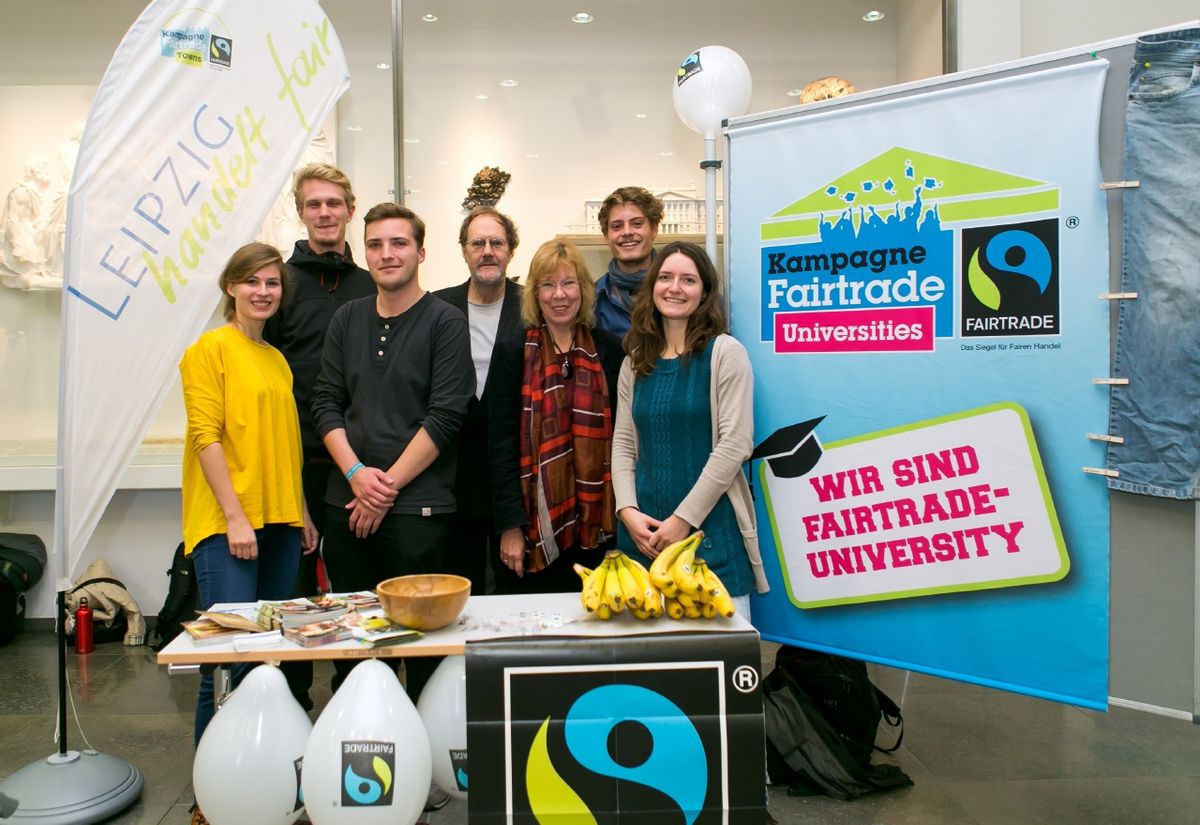 zur Vergrößerungsansicht des Bildes: Foto: Team aus 7 Menschen am Stand mit "Fairtrade University"-Siegel