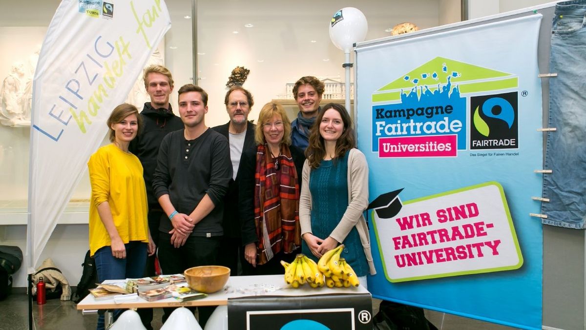 zur Vergrößerungsansicht des Bildes: Foto: Team aus 7 Menschen am Stand mit "Fairtrade University"-Siegel