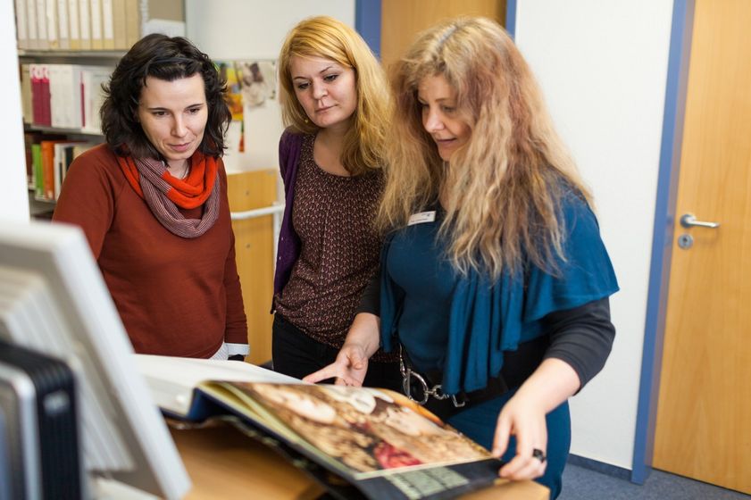 Zwei Studentinnen stehen zusammen mit einer Dozentin in der Kunstbibliothek und schauen sich eine Abbildung in einem Buch an, Foto: Christian Hüller