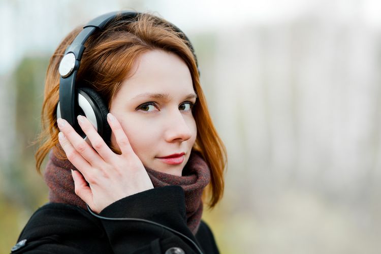 Eine junge Frau im Portrait blickt in die Kamera. Sie hat Kopfhörer auf den Ohren und hält die rechte Hand an ihr Ohr.