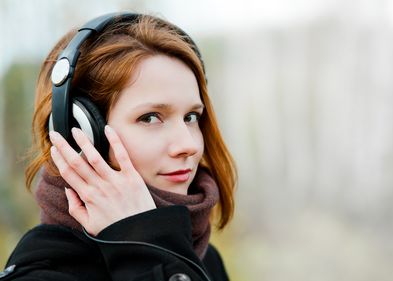 Eine junge Frau im Portrait blickt in die Kamera. Sie hat Kopfhörer auf den Ohren und hält die rechte Hand an ihr Ohr.