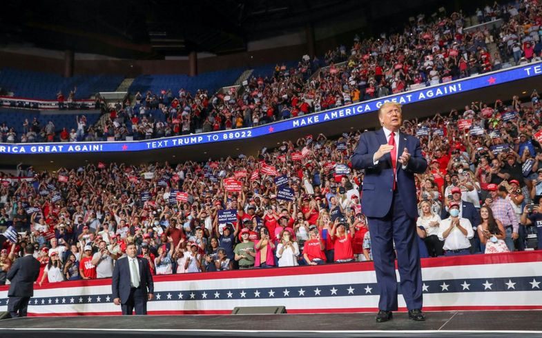 Donald Trump klatscht auf der Bühne bei einer großen Kundgebung in einer Halle, umgeben von jubelnden Unterstützer:innen mit Schildern und Kleidung seiner Kampagne.