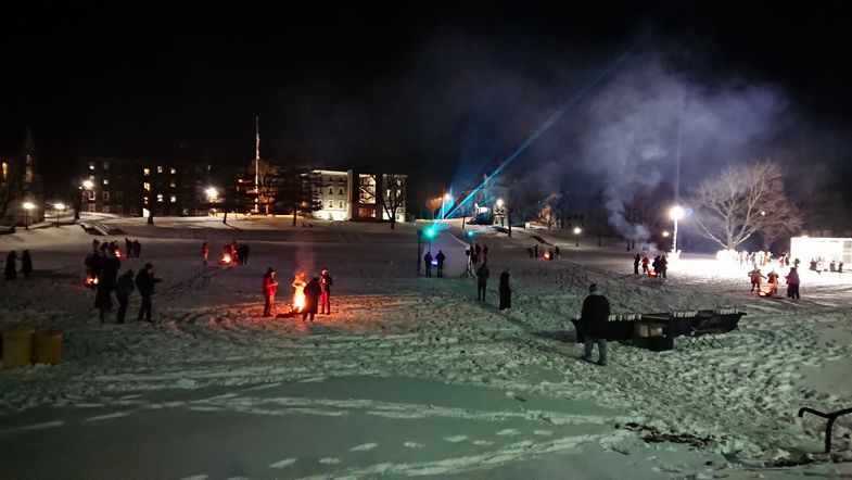 Auf einer zugeschneiten Wiese stehen vereinzelt Menschengruppen um Feuerstellen herum. Es ist dunkel. 
