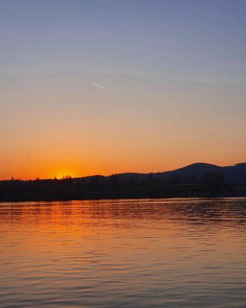 Über der Donau geht die Sonne unter. Im Hintergrund ist ein kleiner Berg zu sehen. Dieser ist schwarz. Der Sonnenuntergang taucht das Bild ansonsten in organgene Farbtöne.