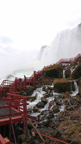 Im Hintergrund des Bildes schießt ein Wasserfall herunter. Weiter vorne im Bild geht eine rote Metalltreppe neben dem Wasserfall empor.