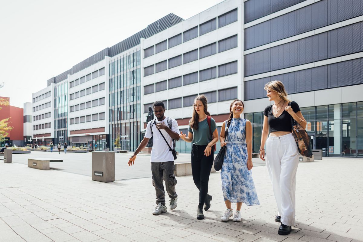 zur Vergrößerungsansicht des Bildes: Colour photo: Four people walking across the inner courtyard of the university and talking