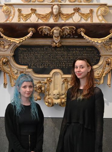 zwei junge Frauen, die die Führung im Rahmen des Wave Gotik Treffen anbieten, stehen vor einem Schnitzepitaph, direkt hinter ihnen ein geschnitzter Totenkopf, das Epitaph ist Weiß und Gold gefassttt