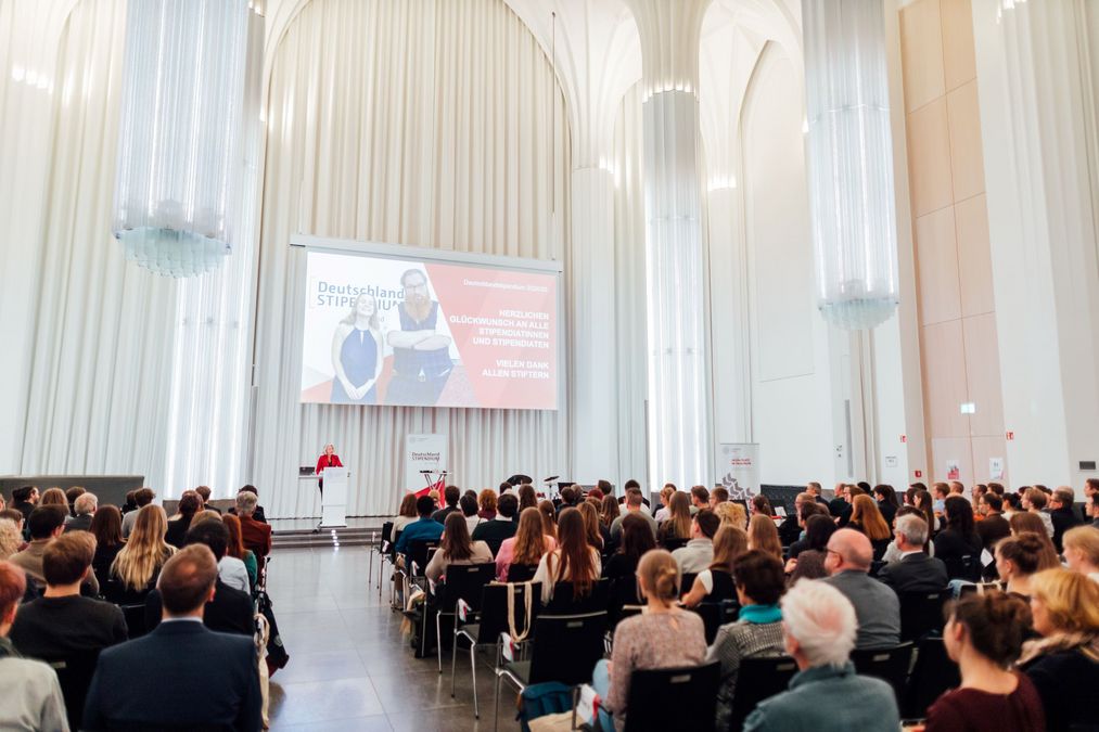 zur Vergrößerungsansicht des Bildes: Förderer und Stipendiaten sitzen im Paulinum und hören der Rede der Rektorin zu, die auf der Bühne zu den Gästen spricht