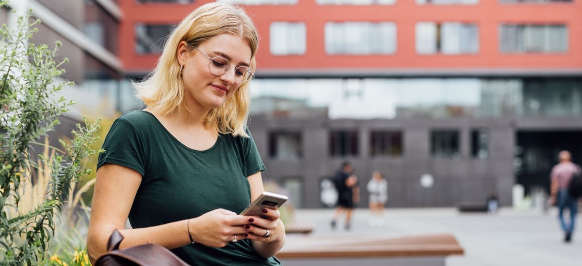Studentin sitzt auf einer Bank auf dem Campus neben ihr steht ihre Tasche und sie schaut auf ihr Handy, Foto: Christian Hüller