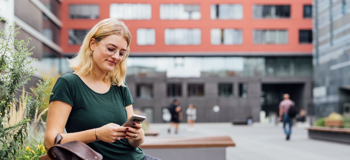 Studentin sitzt auf einer Bank auf dem Campus neben ihr steht ihre Tasche und sie schaut auf ihr Handy, Foto: Christian Hüller