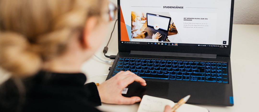 Farbfoto: Eine junge Frau sitzt mit dem Rücken zur Kamer vor einem Laptop und recherchiert auf der Website der Universität Leipzig nach passenden Studiengängen.