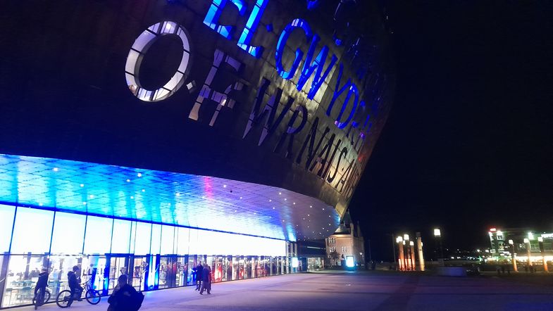 Blick von seitlich-vorn auf das grell beleuchtete Millenium Centre bei Nacht.