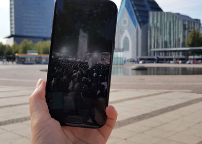 Smartphone zeigt Foto von der Friedlichen Revolution in Leipzig 1989, im Hintergrund das Paulinum