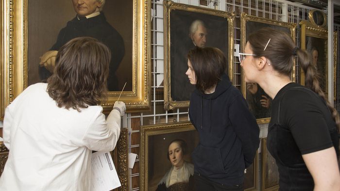 Eine Frau zeigt zwei Studentinnen ein Gemälde.