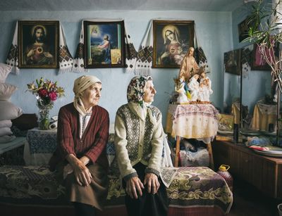 Zu sehen sind zwei ältere Frauen in einem teppicbehangenen Wohnzimmer, die zum Fenster schauen.