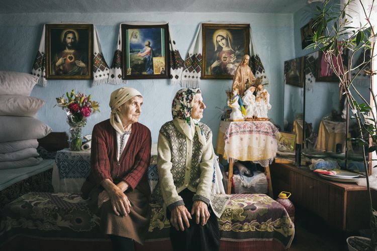 Zu sehen sind zwei ältere Frauen in einem teppicbehangenen Wohnzimmer, die zum Fenster schauen.