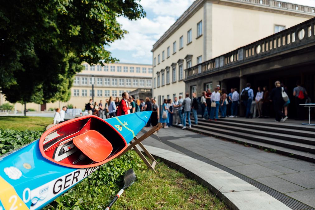 zur Vergrößerungsansicht des Bildes: im Vordergrund ist das hellblau-rote Kajak einer Stipendiatin, im Hintergrund stehen Fördernde und Stipendiaten und unterhalten sich