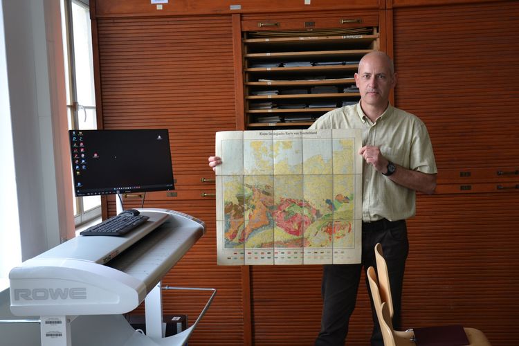 Auf dem Bild ist Gernot Decker zu sehen. Er zeigt eine Karte aus dem Bestand der geowissenschaftlichen Kartensammlung, links im Bild der Großformatscanner. 
