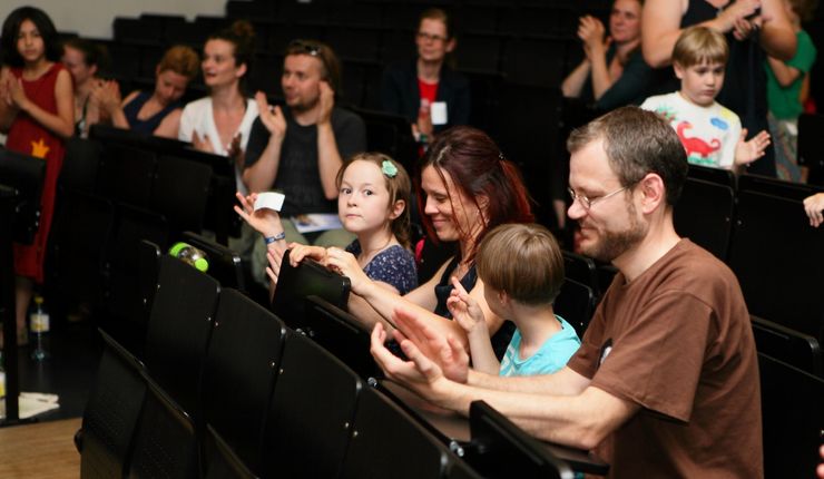 In einem Hörsaal sitzen erwachsene und Kinder in einer Reihe, die gemeinsam einer Vorlesung folgen.