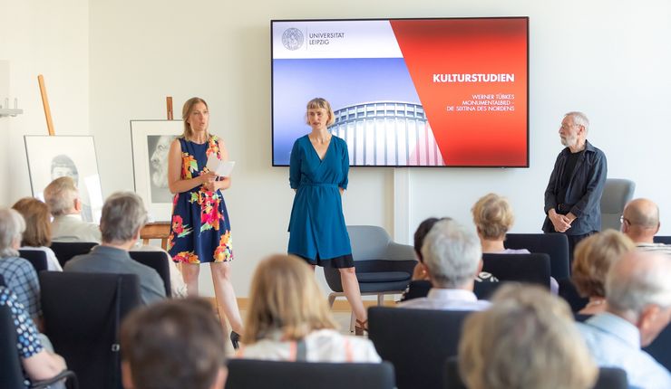 Blick in einen Seminarraum zu einer Veranstaltung der Kulturstudien. Im Vordergrund Blick über das Publikum hinweg, im Hintergrund Gesprächs- und Vortragssituation mit drei Personen