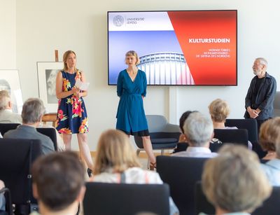 Blick in einen Seminarraum zu einer Veranstaltung der Kulturstudien. Im Vordergrund Blick über das Publikum hinweg, im Hintergrund Gesprächs- und Vortragssituation mit drei Personen