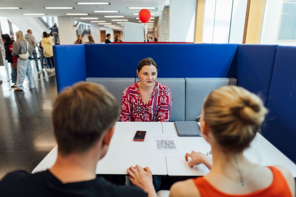 zur Vergrößerungsansicht des Bildes: Foto: Zwei Besucher:innen im Gespräch mit einer Lehramtsstudentin.