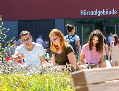Junge Menschen sitzen im Innenhof des Campus Augustusplatz in der Sonne und sprechen miteinander oder lesen. Im Vordergrund sind noch die Grünpflanzen an den Bänken zu sehen.