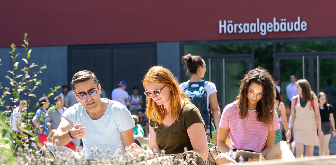 Junge Menschen sitzen im Innenhof des Campus Augustusplatz in der Sonne und sprechen miteinander oder lesen. Im Vordergrund sind noch die Grünpflanzen an den Bänken zu sehen.