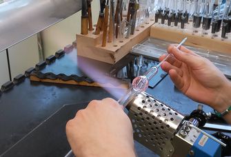 Zu sehen ist, wie ein Apparat aus Glas über der blauen Wasserstoffflamme gehalten wird, damit das Glas geformt werden kann.