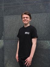 der Deutschlandstipendiat Mika Wilsky trägt schwarze Kleidung und steht vor einer grauen Steinwand