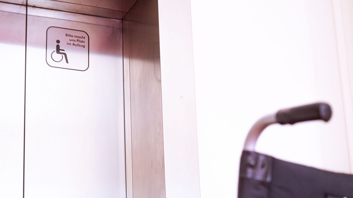 Leere Rollstuhl steht vor einer geschlossen Fahrstuhltür, auf der Tür ist ein Aufkleber mit einem Rollstuhl Piktogramm und den Text "Bitte macht uns Platz im Aufzug)