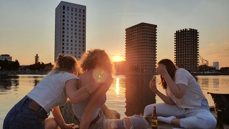 Farbfoto: Aufnahme von drei Frauen von hinten, die bei Sonnenuntergang am Wasser sitzen. Im Hintegrund befinden sich drei Hochhäuser