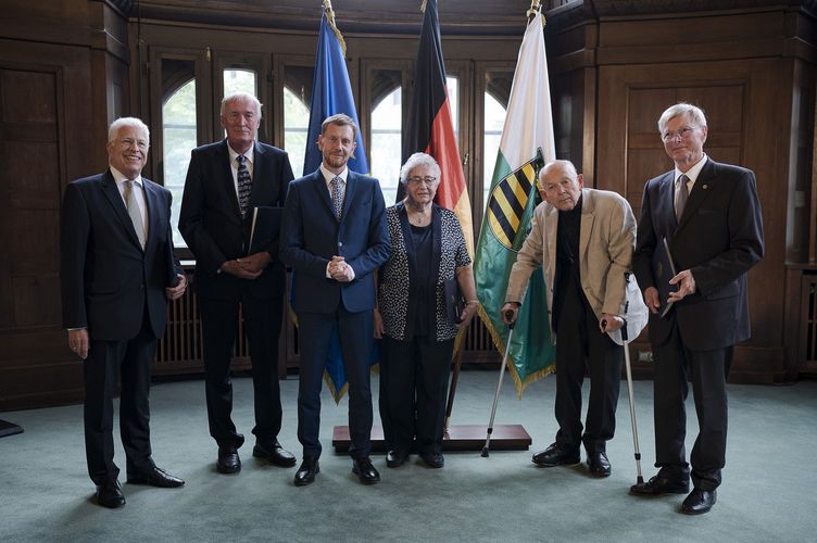 Ministerpräsident Michael Kretschmer in der MItte, geehrte Bürgerinnen und Bürger neben ihm, im Hintergrund Flaggen.