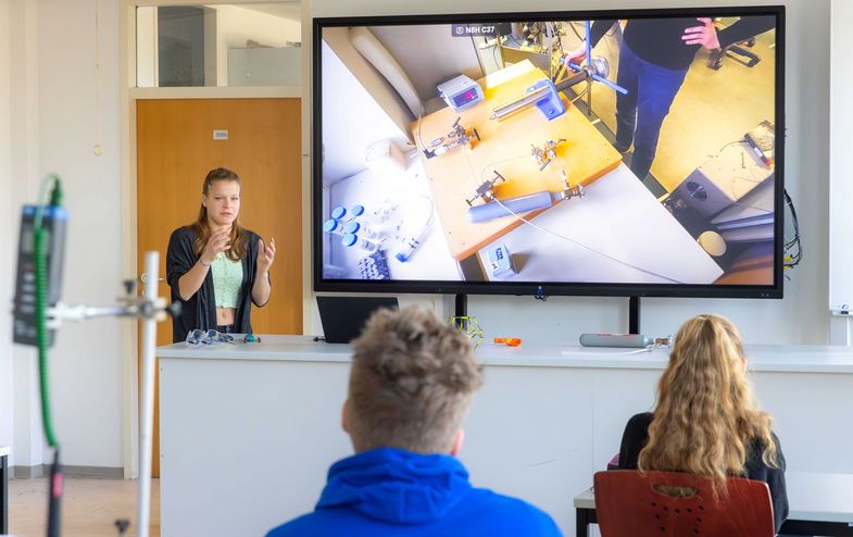 Zu sehen ist, wie Studentin Sindy Krabsch Schüler:innen, vor einemn Monitor, einen Experimentieraufbau zeigt und erklärt. 