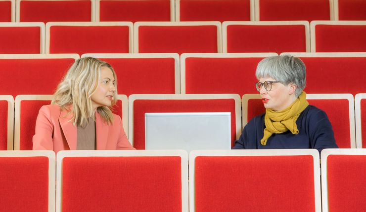 Zwei Frauen unterhalten sich in einem Hörsaal zueinander gewandt. Zwischen Ihnen steht in der Mitte ein Laptop.