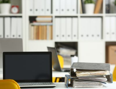 Frontalansicht auf einen Schreibtisch mit Laptop im Vordergrund, im Hintergrund ein Regal mit Büchern und sehr vielen Aktenordnern