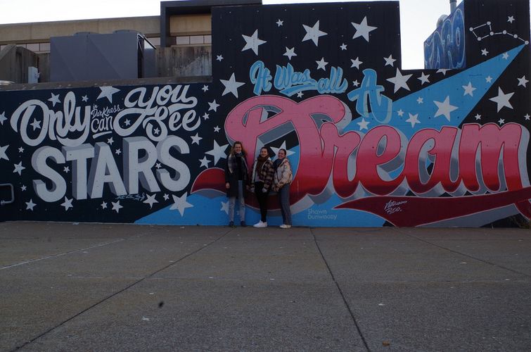Drei Frauen stehen vor einer Wand, welche ein Graffiti trägt. Auf der Wand steht "It was all a dream".