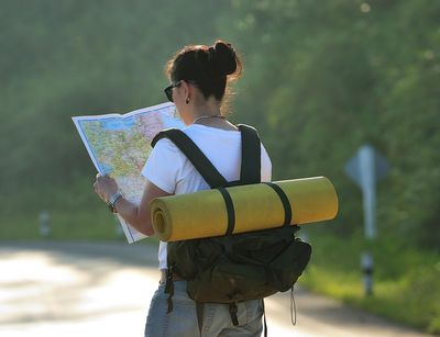 Person steht mit dem Rücken zur Kamera, trägt einen Rucksack, an welchem eine gelbe Isomatte befestigt ist und schaut auf eine Landkarte. Im Hintergrund ist eine begrünte Straße zu sehen.