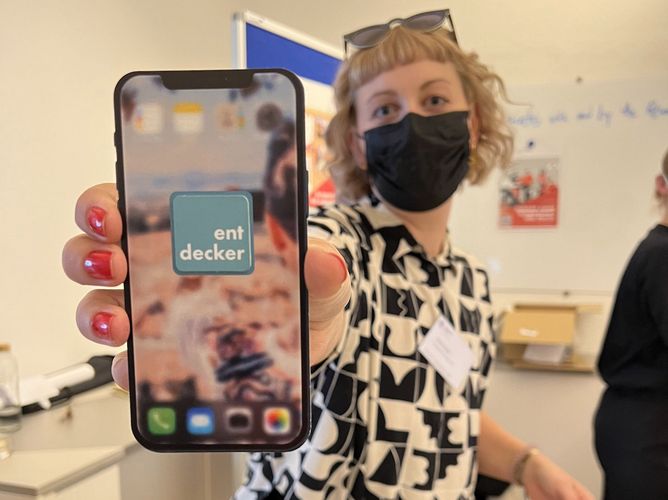 Eine Person hält ein Mobiltelefon hoch, auf dem Display ist der Schriftzug "Entdecker" zu sehen, der für die Entdecker-App der Stabsstelle internationales steht.