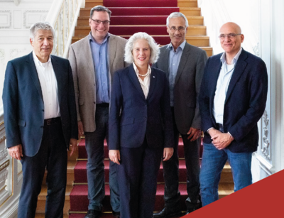 Farbfotografie: Gruppenfoto auf einer Treppe im historischen Rektoratsgebäude. Die Frau steht in der Mitte, rechts und links stehen je zwei Männer. Im unteren Bereich des Bildes sind zwei weinrote Farbflächen und die Aufschrift "Jahresbericht 2022" mit weißen Buchstaben zu sehen.