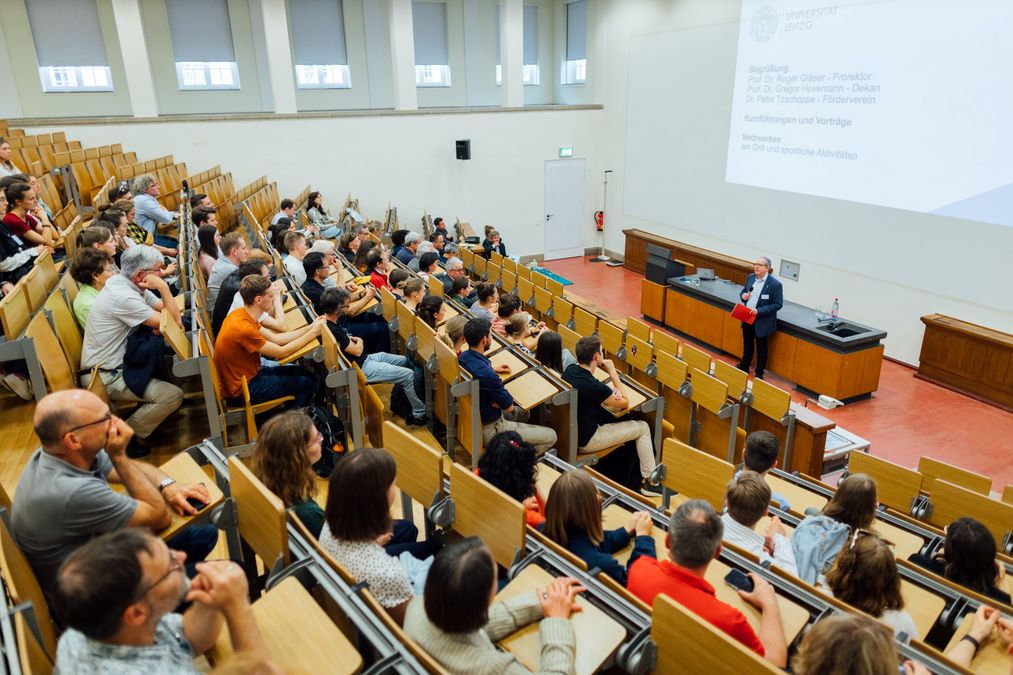 zur Vergrößerungsansicht des Bildes: Studierende und Fördernde sitzen in Hörsaal, vorn steht Professor Gläser und begrüßt die Gäste zum Sommerfest, an der Wand ist ein Folie mit den Informationen zum Sommerfest eingeblendet