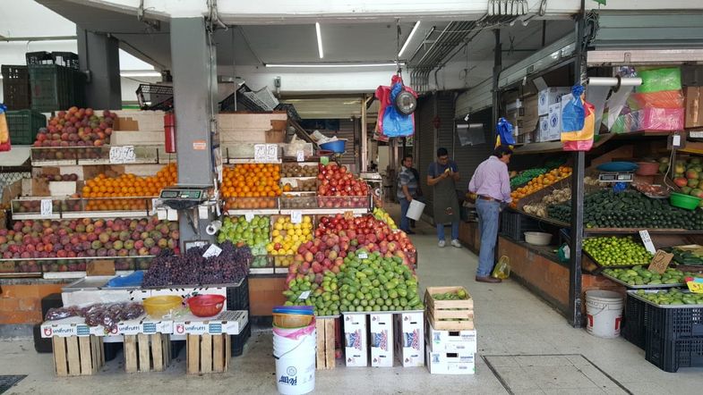 In einer Markthalle ist ein Marktstand zu sehen, welcher vollgepackt mit buntem Obst und Gemüse ist.