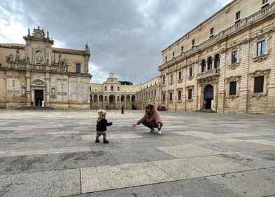 Ein Kleinkind läuft auf einem weitläufigen Platz seiner Mutter entgegen, die in der Hocke ist und ihre Arme dem Kind entgegenstreckt. Der Platz ist umsäumt von historischen Gebäuden. Im Hintergrund sieht man vereinzelt Menschen. 