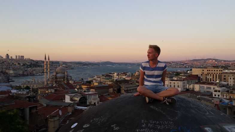 Eine Person sitzt auf einer Kugel und schaut nach links. Hinter der Person ist Istanbul zu sehen. Die Meerenge Bosporus schlängelt sich durch die Stadt. Die Sonne geht über der Stadt unter.