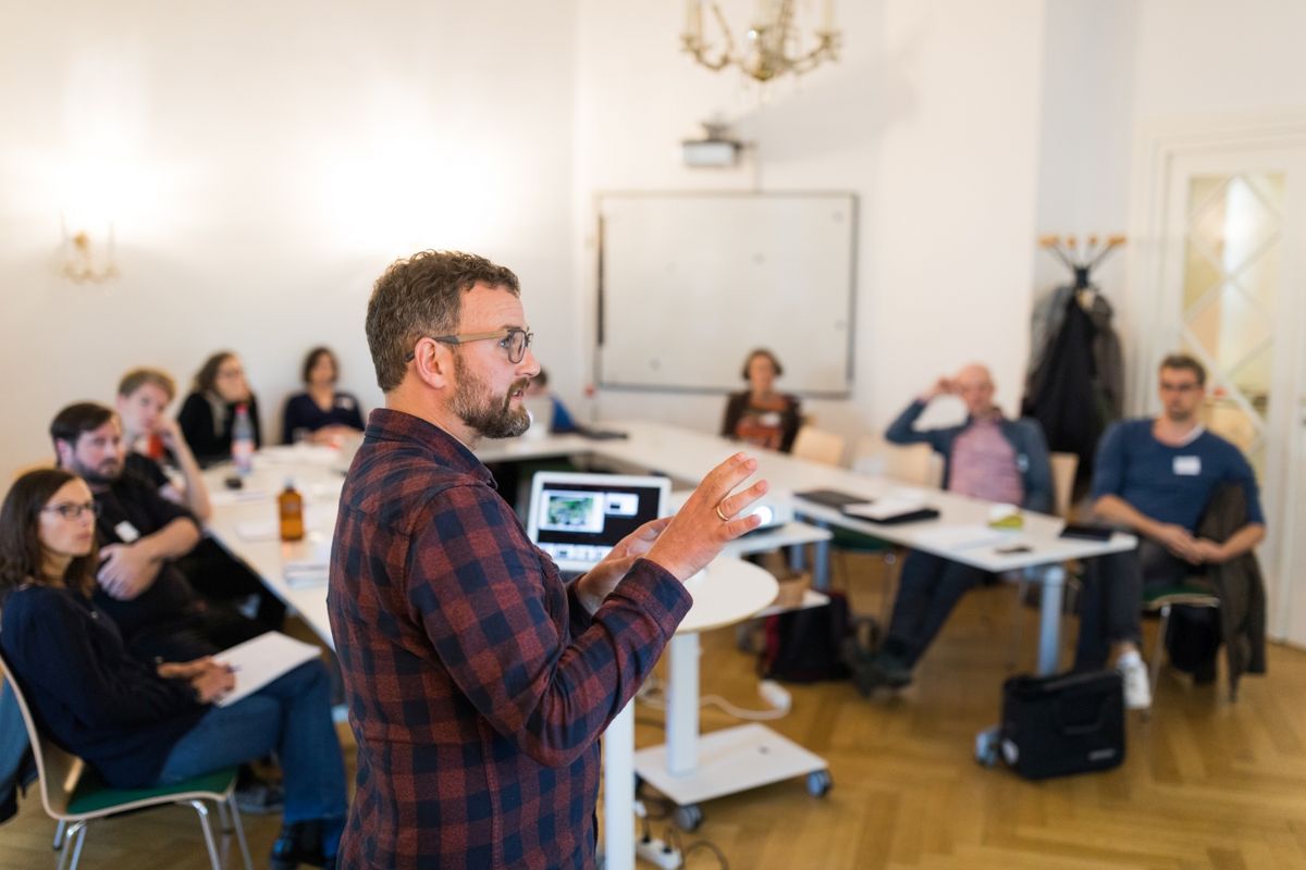 zur Vergrößerungsansicht des Bildes: In einem Seminarraum sitzen viele Personen an einem U-Förmig aufgestellten Tisch. Sieh hören einer weiteren Person zu, die einen Vortrag hält.